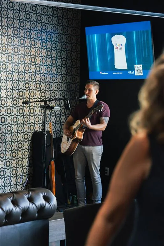 Man singing and playing acoustic guitar on stage with a TV screen behind him